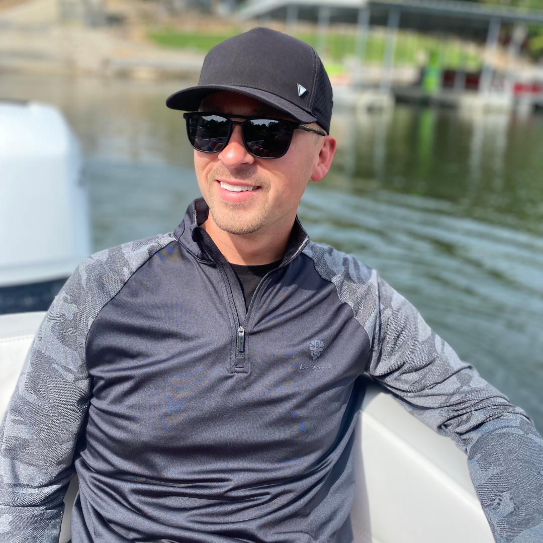 Man wearing sunglasses and a cap on a boat by a body of water